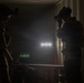 Two military or tactical personnel are silhouetted in a dark, enclosed space that appears to be a shipping container or similar structure. One individual is raising an optical device (possibly binoculars or a camera) to their face, while both wear tactical helmets with mounted equipment. Bright artificial lights and hazy atmospheric conditions are visible in the background.