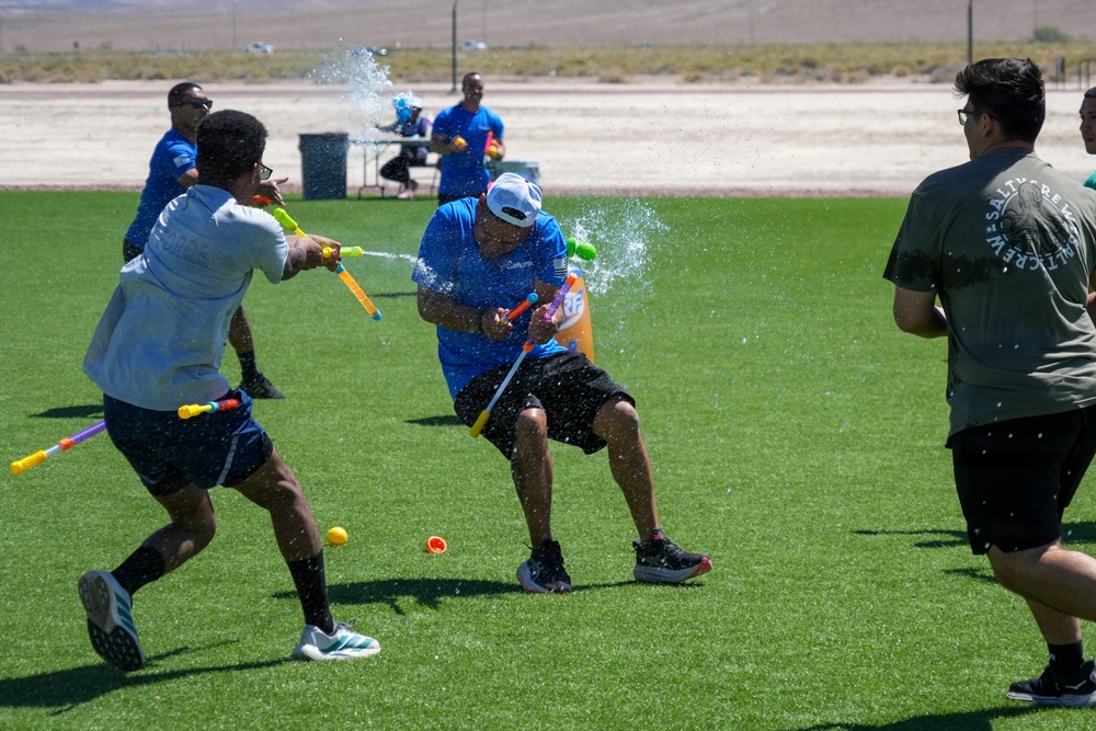 A group of people are engaged in an outdoor water gun fight on an artificial turf field. The participants are using colorful water guns and water balloons, with water visibly spraying in multiple directions. The event appears to be a casual recreational or team-building activity taking place in a sunny, arid environment.