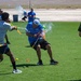 A group of people are engaged in an outdoor water gun fight on an artificial turf field. The participants are using colorful water guns and water balloons, with water visibly spraying in multiple directions. The event appears to be a casual recreational or team-building activity taking place in a sunny, arid environment.
