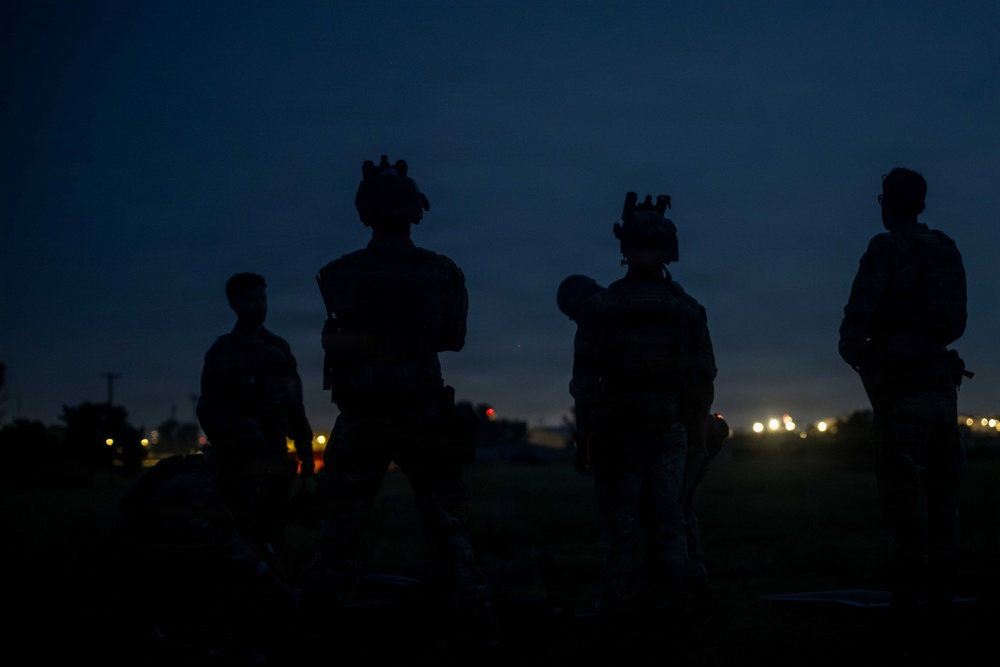 A nighttime photograph showing four silhouetted figures in tactical gear standing in an outdoor area. The figures appear to be equipped with helmets, body armor, and tactical equipment. Distant lights illuminate the dark horizon behind them.