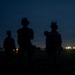 A nighttime photograph showing four silhouetted figures in tactical gear standing in an outdoor area. The figures appear to be equipped with helmets, body armor, and tactical equipment. Distant lights illuminate the dark horizon behind them.
