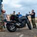 A motorcycle safety or rider training course is taking place in an outdoor parking area, likely on a military installation. An instructor in civilian clothes is demonstrating motorcycle components to a group of military personnel and civilians gathered around a black sport motorcycle. A helmet and gear bag are visible on the motorcycle seat, suggesting a hands-on training session.