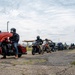 A group of motorcyclists are lined up in a parking lot or staging area, appearing to prepare for a group ride. The riders are wearing helmets and riding gear, with a variety of motorcycle styles visible including a prominent red Indian motorcycle in the foreground. The setting appears to be near a fenced facility under an overcast sky.