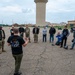 A group of approximately 15 people are gathered in a large paved area, likely on a military installation, listening to a briefing from a man in a black t-shirt facing away from the camera. Several motorcycles are visible in the background, and some participants are wearing military uniforms while others are in civilian motorcycle gear. A large water tower and military-style buildings are visible in the background.