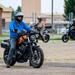 A group of motorcyclists are participating in what appears to be a motorcycle safety or training course in a paved lot near a military or institutional facility. The foreground rider, wearing a blue jacket and full-face helmet, is maneuvering an orange and black cruiser-style motorcycle. Several other riders on various motorcycle types are visible in the background, some in military-style clothing.