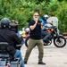 A motorcycle training or instruction session taking place in an open paved area. A standing instructor with a white beard, wearing a black t-shirt and olive pants, is gesturing and communicating with two motorcycle riders. The setting appears to be an informal training ground with green vegetation in the background.