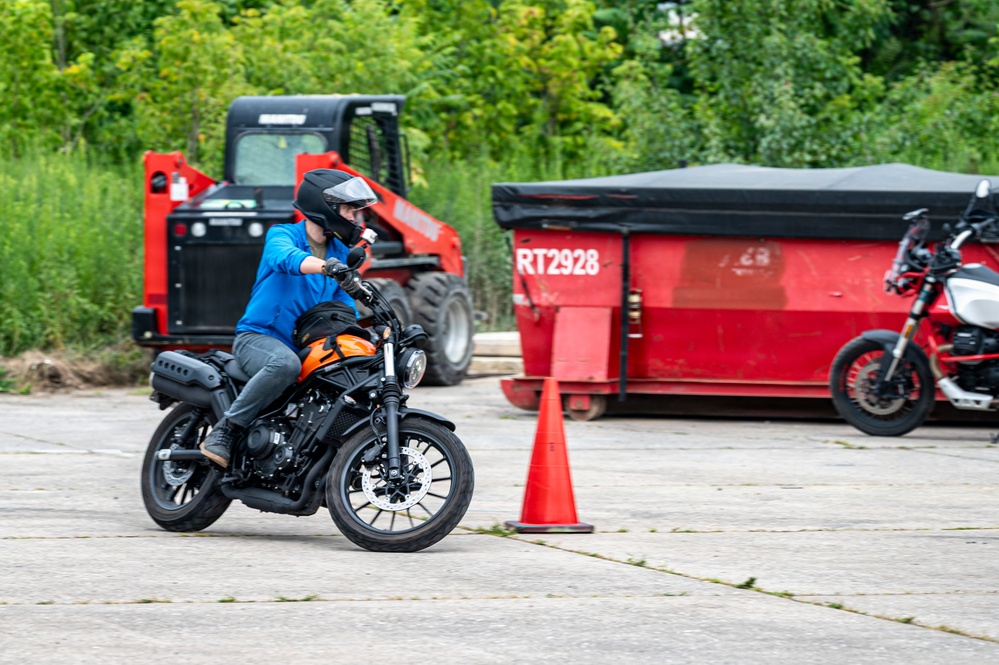 A motorcyclist wearing a blue hoodie, gray pants, and a black helmet is riding an orange and black motorcycle on a paved lot, navigating around an orange traffic cone. The setting appears to be a motorcycle training or practice course, with a red skid steer loader and a red dumpster container labeled 'RT2928' visible in the background. A second motorcycle is partially visible on the far right of the frame.