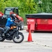 A motorcyclist wearing a blue hoodie, gray pants, and a black helmet is riding an orange and black motorcycle on a paved lot, navigating around an orange traffic cone. The setting appears to be a motorcycle training or practice course, with a red skid steer loader and a red dumpster container labeled 'RT2928' visible in the background. A second motorcycle is partially visible on the far right of the frame.