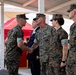 A formal U.S. Marine Corps military ceremony is taking place under a covered outdoor pavilion. Two Marines are shaking hands in what appears to be a receiving line or award/recognition event, with other uniformed Marines and a civilian in a suit standing nearby. An audience is visible in the background near a red tent structure.