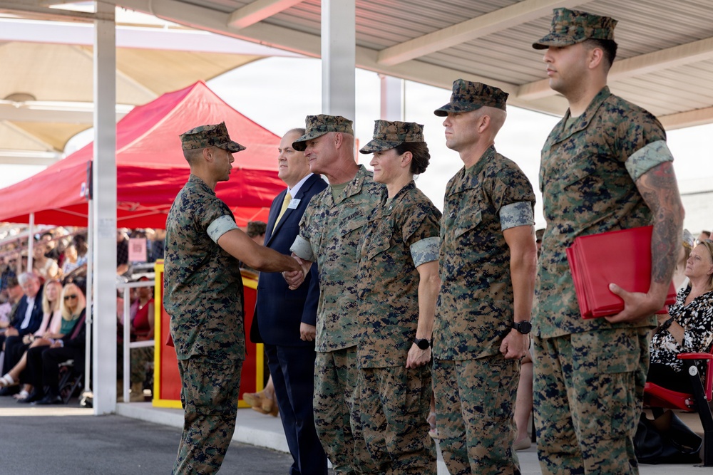 A formal U.S. Marine Corps military ceremony is taking place under a covered outdoor pavilion. A Marine on the left is shaking hands with a senior officer in a receiving line, while other uniformed Marines and a civilian in a suit stand in formation. An audience of civilians and military personnel is visible in the background near a red tent.