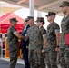 A formal U.S. Marine Corps military ceremony is taking place under a covered outdoor pavilion. A Marine on the left is shaking hands with a senior officer in a receiving line, while other uniformed Marines and a civilian in a suit stand in formation. An audience of civilians and military personnel is visible in the background near a red tent.