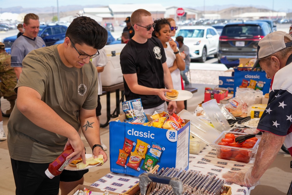An outdoor food service event, likely a military appreciation barbecue or community cookout, taking place in a parking lot. People are lined up at a food table stocked with chips, condiments, and burger fixings. An older volunteer wearing an American flag shirt is serving food to attendees.