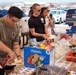 An outdoor food service event, likely a military appreciation barbecue or community cookout, taking place in a parking lot. People are lined up at a food table stocked with chips, condiments, and burger fixings. An older volunteer wearing an American flag shirt is serving food to attendees.