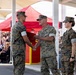 A military ceremony is taking place outdoors, likely at a U.S. Marine Corps installation, where a senior officer is shaking hands with a junior Marine while passing a folded flag or document. Two additional Marines stand at attention in the background, and a seated audience of civilians and military personnel observes the event. The setting and uniforms indicate a formal ceremony such as a promotion, retirement, or change of command.
