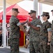 A formal military ceremony is taking place outdoors, likely an award or recognition presentation. Two Marines in the foreground are shaking hands while one passes a red document or certificate to the other. Additional uniformed personnel stand at attention in the background, with a seated audience visible under a covered structure.