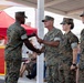 A military ceremony is taking place under a covered outdoor pavilion, where a service member is receiving a document and handshake from a senior officer. Four personnel in U.S. Marine Corps MARPAT woodland camouflage uniforms are visible in the foreground, with an audience of civilians seated in the background. The setting appears to be a formal recognition or award ceremony.