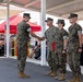 A military ceremony is taking place outdoors under a covered structure, where a service member is shaking hands with a senior officer in a line of uniformed personnel. The setting appears to be a formal recognition or award ceremony, with an audience of civilians and military personnel seated in bleachers in the background. All individuals are wearing U.S. Marine Corps MARPAT woodland camouflage uniforms.