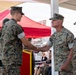A formal U.S. Marine Corps ceremony is taking place outdoors, where a senior Marine officer is shaking hands with a junior Marine, likely presenting an award or recognition. The recipient is holding what appears to be a certificate or award case. A third Marine, appearing to be female, stands nearby observing the exchange.