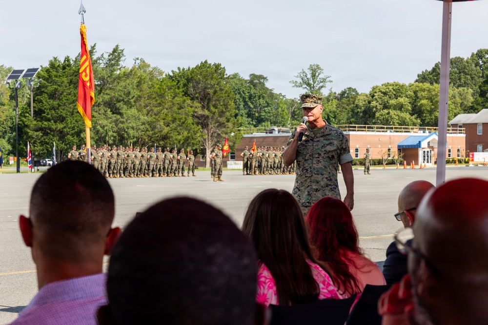A U.S. Marine Corps officer in camouflage utility uniform is addressing an audience via microphone during what appears to be a formal military ceremony on a parade deck. Behind him, a large formation of Marines in uniform stands at attention, with a red and gold Marine Corps guidon flag prominently displayed. The setting appears to be a military installation with brick buildings and trees visible in the background.