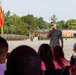 A U.S. Marine Corps officer in camouflage utility uniform is addressing an audience via microphone during what appears to be a formal military ceremony on a parade deck. Behind him, a large formation of Marines in uniform stands at attention, with a red and gold Marine Corps guidon flag prominently displayed. The setting appears to be a military installation with brick buildings and trees visible in the background.