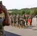A formal U.S. Marine Corps parade or pass-in-review ceremony is taking place on a paved parade deck. Marines in woodland MARPAT camouflage uniforms are marching in formation while carrying a red unit guidon. A senior Marine in the foreground renders a salute as the formation passes.