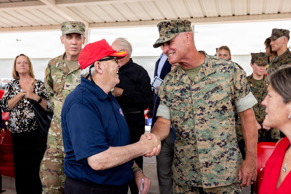 A military greeting ceremony is taking place under a covered outdoor structure. A U.S. Marine officer in MARPAT digital camouflage is shaking hands with an elderly civilian wearing a red cap and navy blue polo shirt. Multiple military personnel from different branches and civilians are present in the background.