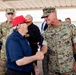 A military greeting ceremony is taking place under a covered outdoor structure. A U.S. Marine officer in MARPAT digital camouflage is shaking hands with an elderly civilian wearing a red cap and navy blue polo shirt. Multiple military personnel from different branches and civilians are present in the background.