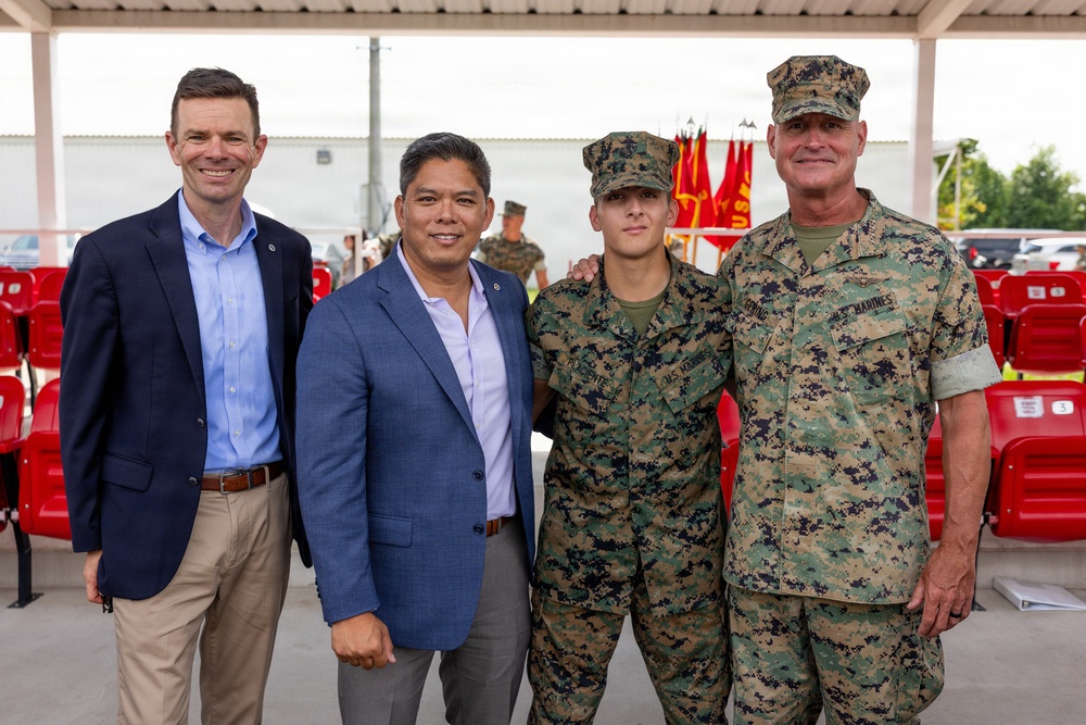 Four individuals pose for a group photo at what appears to be a U.S. Marine Corps ceremony or event. Two men in civilian business attire stand alongside two individuals in U.S. Marine Corps MARPAT camouflage uniforms. The setting includes red stadium-style seating and Marine Corps flags visible in the background.