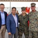 Four individuals pose for a group photo at what appears to be a U.S. Marine Corps ceremony or event. Two men in civilian business attire stand alongside two individuals in U.S. Marine Corps MARPAT camouflage uniforms. The setting includes red stadium-style seating and Marine Corps flags visible in the background.