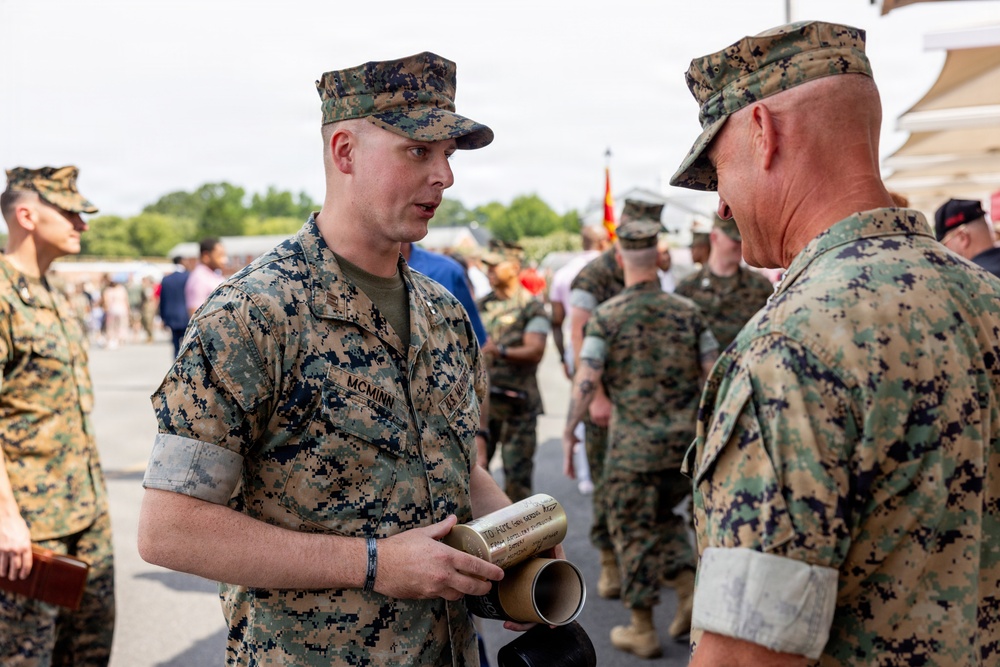 Two U.S. Marines in MARPAT woodland camouflage uniforms are engaged in conversation at what appears to be a military ceremony or event. The Marine facing the camera, identified by his name tape as 'McMinn,' is holding a cylindrical brass or gold-colored object, possibly an artillery shell casing or commemorative item with engraved text. A crowd of military personnel and civilians is visible in the background.