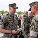 Two U.S. Marines in MARPAT woodland camouflage uniforms are engaged in conversation at what appears to be a military ceremony or event. The Marine facing the camera, identified by his name tape as 'McMinn,' is holding a cylindrical brass or gold-colored object, possibly an artillery shell casing or commemorative item with engraved text. A crowd of military personnel and civilians is visible in the background.