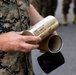 A person in U.S. Marine Corps MARPAT digital camouflage uniform is holding a brass artillery shell casing with handwritten inscriptions on it. The casing appears to be a commemorative or presentation item signed by multiple personnel from an Artillery Battery. A cardboard tube and black cap are also visible, suggesting the casing was stored or transported in a protective container.