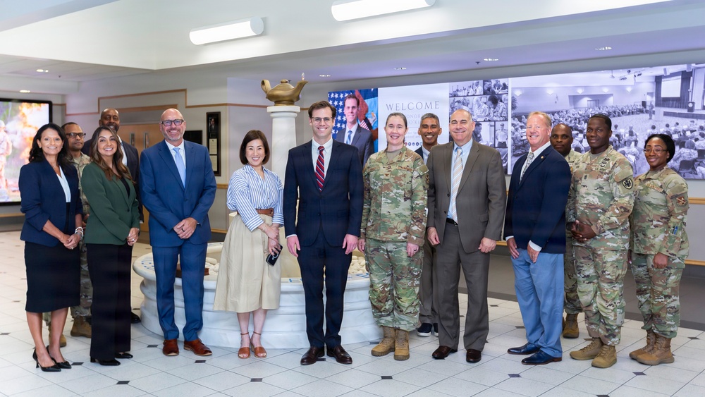 A group of approximately 14 individuals poses for a formal group photograph in what appears to be a military installation lobby or reception area. The group includes a mix of uniformed military personnel in Army Combat Uniforms (ACU/OCP pattern) and civilians in business attire. A 'WELCOME' banner is visible on a display screen in the background, suggesting an official visit or ceremony.
