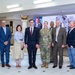 A group of approximately 14 individuals poses for a formal group photograph in what appears to be a military installation lobby or reception area. The group includes a mix of uniformed military personnel in Army Combat Uniforms (ACU/OCP pattern) and civilians in business attire. A 'WELCOME' banner is visible on a display screen in the background, suggesting an official visit or ceremony.