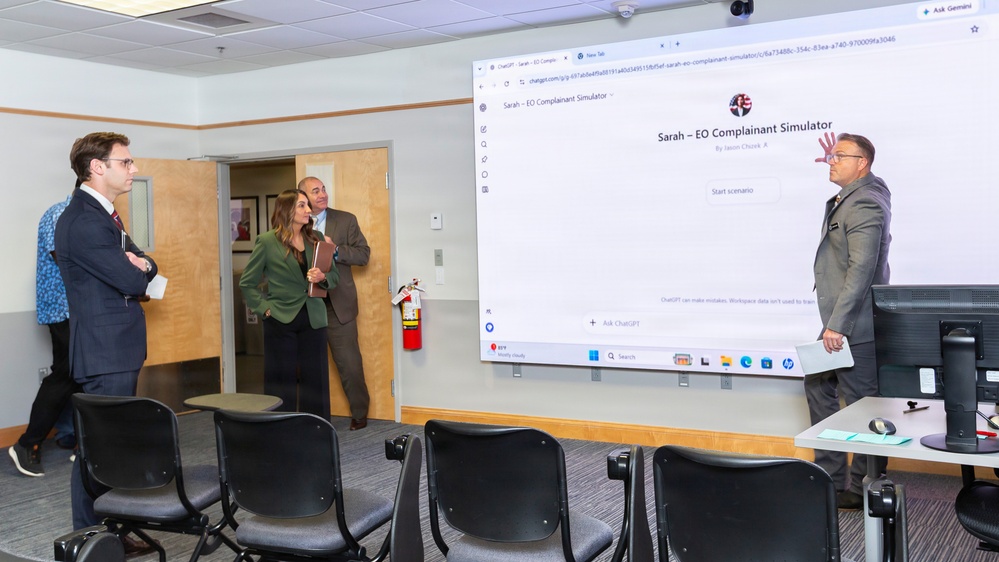 A professional presentation or demonstration is taking place in a conference or training room. A large projected screen displays a ChatGPT-based application called 'Sarah – EO Complainant Simulator' created by Jason Chusek. Four individuals in business attire are gathered, with one person appearing to present near the screen while others observe.