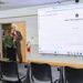 A professional presentation or demonstration is taking place in a conference or training room. A large projected screen displays a ChatGPT-based application called 'Sarah – EO Complainant Simulator' created by Jason Chusek. Four individuals in business attire are gathered, with one person appearing to present near the screen while others observe.
