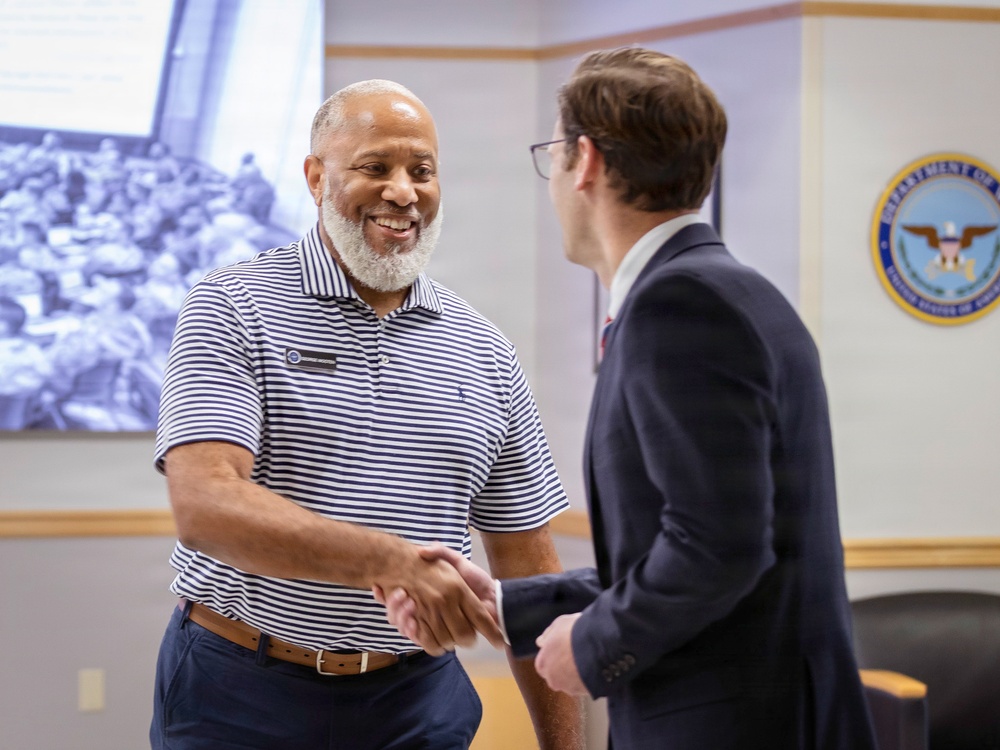 Two men are shaking hands in what appears to be an official government setting, likely a Department of Veterans Affairs facility. The man on the left is smiling broadly, wearing a striped polo shirt with a name badge, while the man on the right is dressed in a dark business suit. The interaction appears cordial and professional in nature.