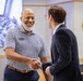 Two men are shaking hands in what appears to be an official government setting, likely a Department of Veterans Affairs facility. The man on the left is smiling broadly, wearing a striped polo shirt with a name badge, while the man on the right is dressed in a dark business suit. The interaction appears cordial and professional in nature.