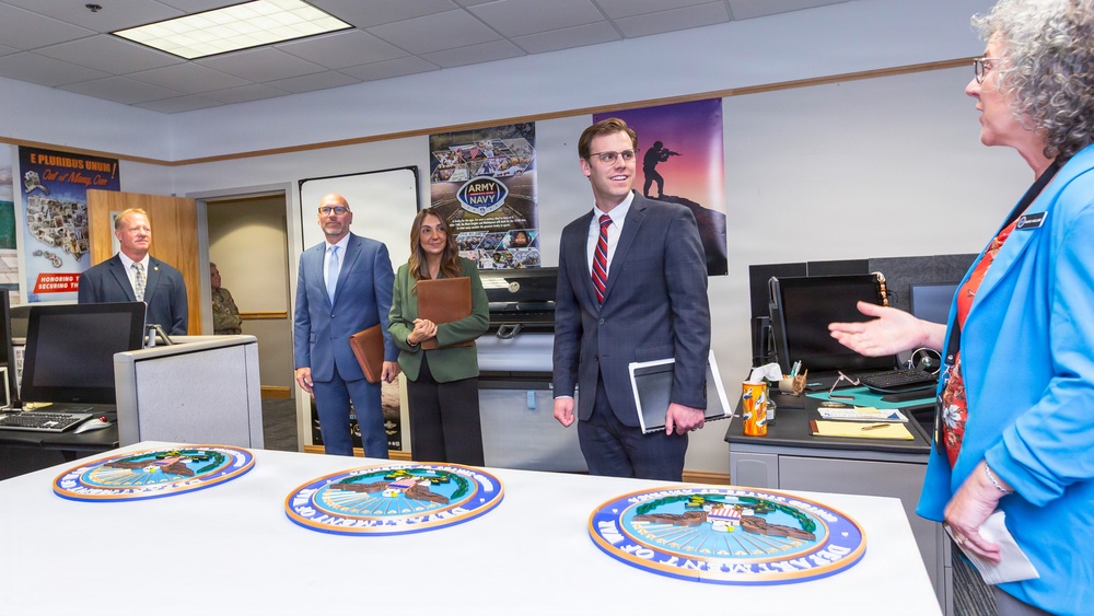 A formal meeting or briefing is taking place in what appears to be a military or government office setting. Several professionally dressed individuals are gathered around a table displaying multiple official seals, while a woman in a blue blazer appears to be presenting or explaining something. The room contains military recruitment posters and official insignia consistent with a U.S. military administrative environment.
