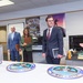 A formal meeting or briefing is taking place in what appears to be a military or government office setting. Several professionally dressed individuals are gathered around a table displaying multiple official seals, while a woman in a blue blazer appears to be presenting or explaining something. The room contains military recruitment posters and official insignia consistent with a U.S. military administrative environment.