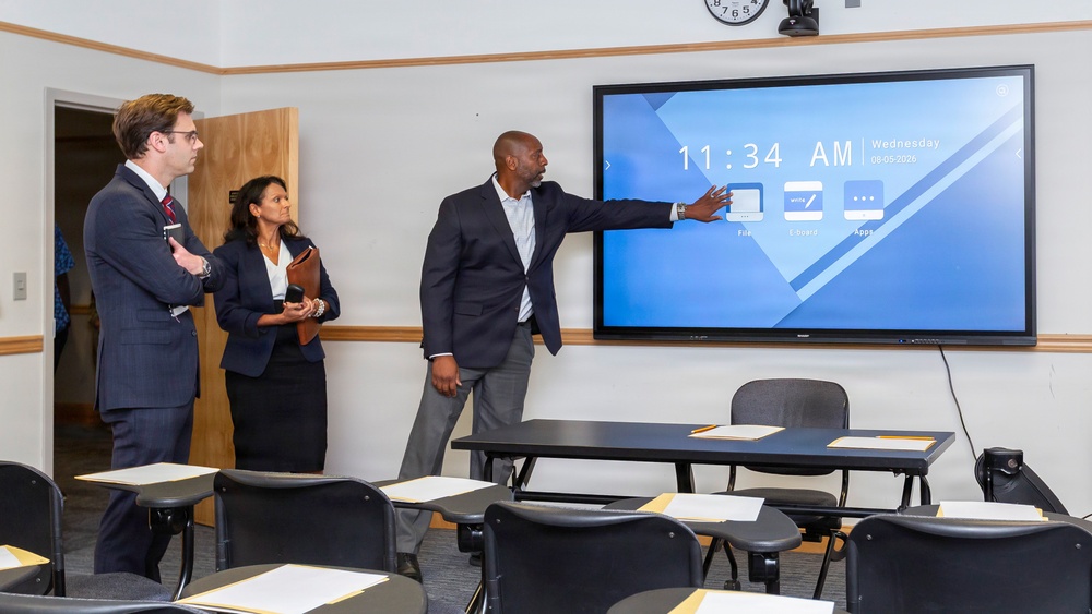 Three professionally dressed individuals are in a classroom or conference room setting. A man is pointing at a large interactive display screen showing the time 11:34 AM on Wednesday, 08-05-2026, apparently demonstrating the technology to two colleagues. The room contains multiple student-style desks with papers arranged on them.