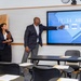 Three professionally dressed individuals are in a classroom or conference room setting. A man is pointing at a large interactive display screen showing the time 11:34 AM on Wednesday, 08-05-2026, apparently demonstrating the technology to two colleagues. The room contains multiple student-style desks with papers arranged on them.