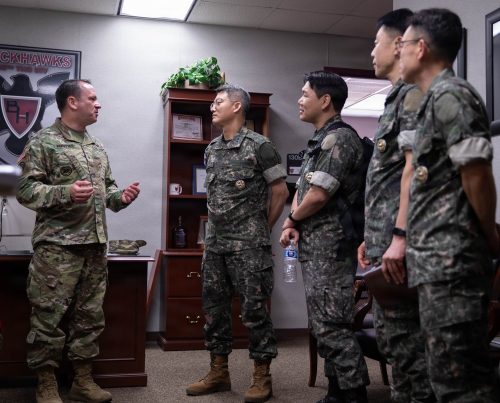 A U.S. Army soldier in OCP (Operational Camouflage Pattern) uniform is briefing or conversing with four military personnel wearing Republic of Korea (ROK) Army uniforms in what appears to be a military office. The setting includes a 'Blackhawks' unit insignia on the wall, suggesting this is a U.S. Army aviation unit headquarters. The interaction appears to be a formal military exchange or liaison visit.