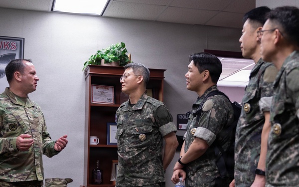 A U.S. Army soldier in OCP (Operational Camouflage Pattern) uniform is briefing or conversing with four military personnel wearing Republic of Korea (ROK) Army uniforms in what appears to be a military office. The setting includes a 'Blackhawks' unit insignia on the wall, suggesting this is a U.S. Army aviation unit headquarters. The interaction appears to be a formal military exchange or liaison visit.