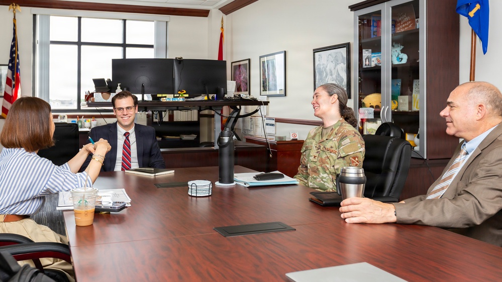 Four individuals are seated around a large conference table in a formal office setting, engaged in what appears to be a professional meeting. One person is wearing a military uniform with camouflage pattern, while the others are in civilian business attire. The atmosphere appears relaxed and collegial, with participants smiling and conversing.