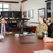 Four individuals are seated around a large conference table in a formal office setting, engaged in what appears to be a professional meeting. One person is wearing a military uniform with camouflage pattern, while the others are in civilian business attire. The atmosphere appears relaxed and collegial, with participants smiling and conversing.