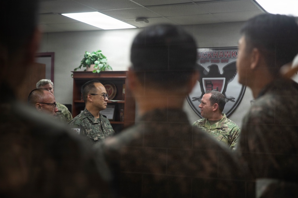 A group of military personnel from what appears to be the U.S. Army and Republic of Korea (ROK) Army are gathered in an indoor office or briefing room. The individuals are engaged in conversation, suggesting a joint meeting, briefing, or exchange visit. A unit insignia on the wall partially reads 'Blackhawks' indicating a U.S. Army aviation or combat unit.