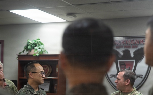 A group of military personnel from what appears to be the U.S. Army and Republic of Korea (ROK) Army are gathered in an indoor office or briefing room. The individuals are engaged in conversation, suggesting a joint meeting, briefing, or exchange visit. A unit insignia on the wall partially reads 'Blackhawks' indicating a U.S. Army aviation or combat unit.