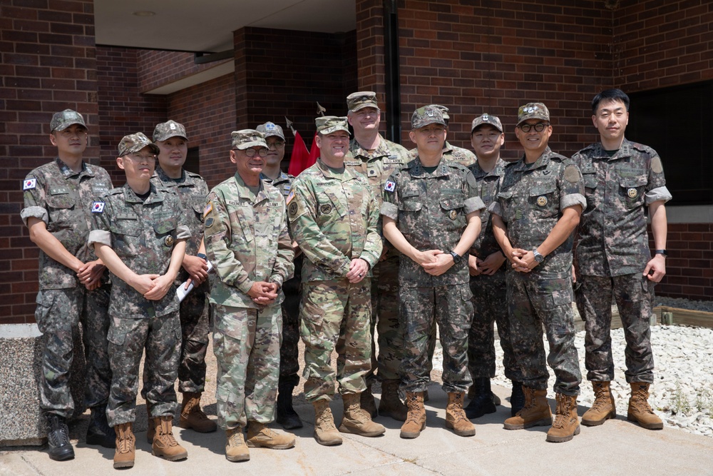 A group of approximately eleven military personnel pose for a formal group photograph in front of a brick building. The group includes both U.S. Army soldiers in OCP (Operational Camouflage Pattern) uniforms and Republic of Korea (ROK) Army soldiers in their distinctive digital camouflage uniforms with South Korean flag patches. This appears to be a joint military exchange or liaison visit between U.S. and South Korean armed forces.