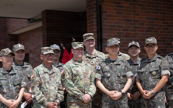 A group of approximately eleven military personnel pose for a formal group photograph in front of a brick building. The group includes both U.S. Army soldiers in OCP (Operational Camouflage Pattern) uniforms and Republic of Korea (ROK) Army soldiers in their distinctive digital camouflage uniforms with South Korean flag patches. This appears to be a joint military exchange or liaison visit between U.S. and South Korean armed forces.